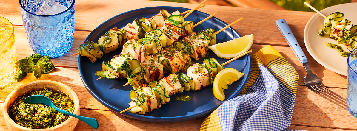 Grilled zucchini ribbon and halloumi skewers with a lemon mint pesto drizzle on a blue plate with lime wedges on the side.