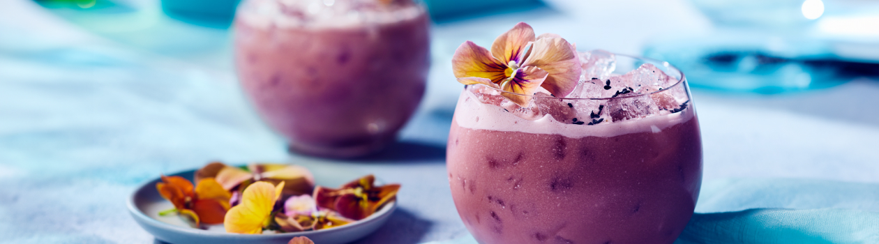 Low rounded glass of purple iced cocktail with edible floral garnish