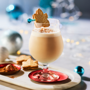 Cookies and Milk cocktail in a bell-shaped short-stemmed glass with a maple crème cookie on a skewer across the rim, all set on a white marble surface with holiday decorations.