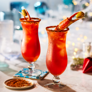 Two tall bell-shaped glasses with a Dirty Caesar cocktail and king crab leg garnish on a white marble surface with holiday decorations in the background.