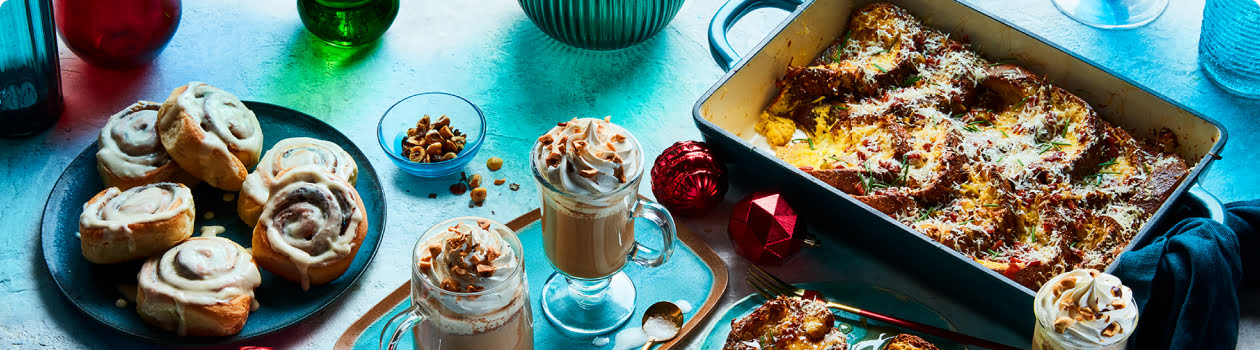 Light blue table surface with a serving plate of glazed cinnamon buns, eggnog french toast, two whip cream topped lattes, and a casserole dish of strata