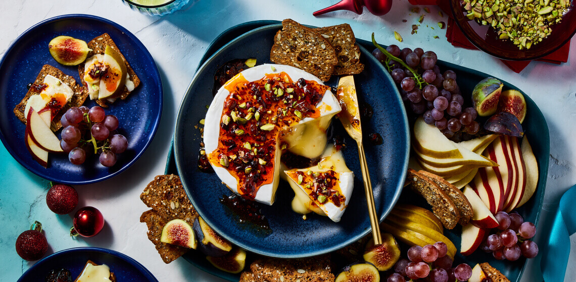 Overhead shot of baked brie topped with hot honey and pistachios with crips, apples, and figs to the side.