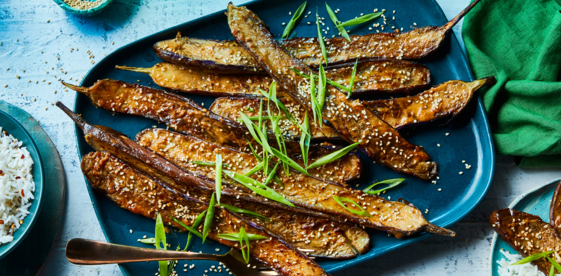 Rectangular blue serving platter with Japanese eggplant that has been roasted and garnished with fresh green onion and toasted sesame seeds with a side of white rice in a bowl