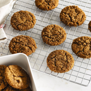 Chewy ginger molasses cookies on a cooling rack and in a white bowl on a white countertop