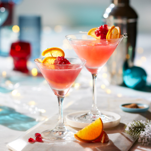 Pom Pomtini in two martini glasses on a white marble surface with holiday decorations and a martini shaker in the background.