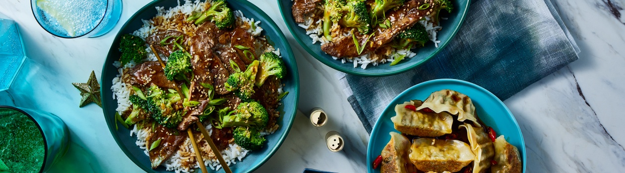 Two plates of beef and broccoli on a bed of rice with a side of potstickers