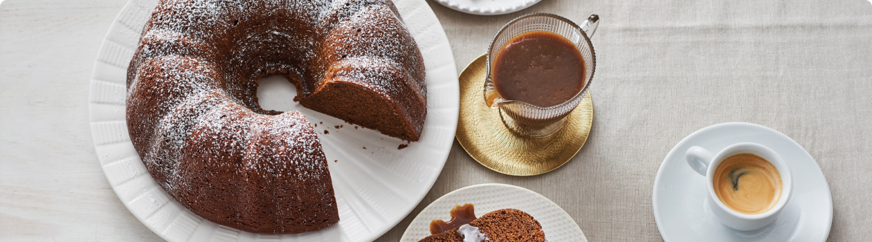 Gingerbread Bundt Cake sitting on a white cake plate, dusted with icing sugar and next to a container full of Coffee Caramel Sauce.