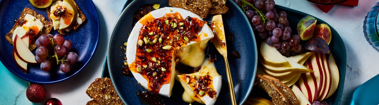 Overhead shot of baked brie topped with hot honey and pistachios with crips, apples, and figs to the side.