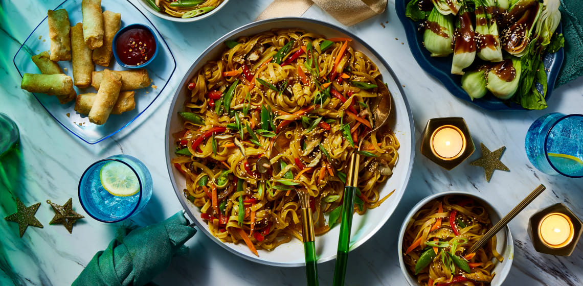 Bowl of vegetable Singapore noodles with a side of spring rolls and bok choy.