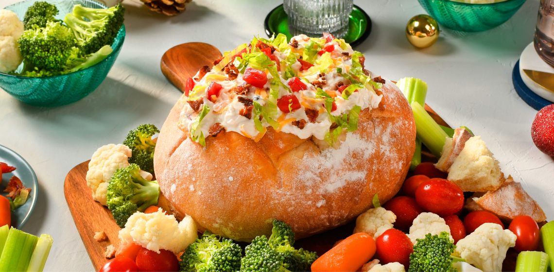 BLT dip in a bread bowl surrounded by a mix of vegetables on a chopping board