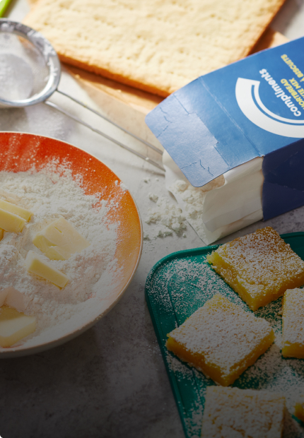 Marble surface with box of Compliments Shortbread cookie mix, baked lemon squares and a mixing bowl of cookie mix and butter.