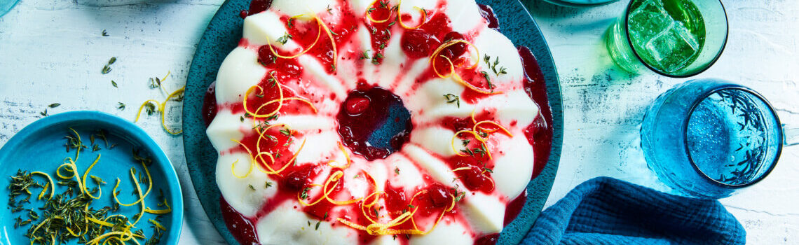 Round blue cake platter with chilled cranberry-lemon blancmange, with cranberry sauce, lemon peel and thyme leaf garnish.