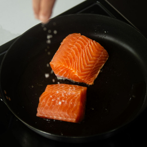 Two raw salmon fillets in a pan being sprinkled with salt