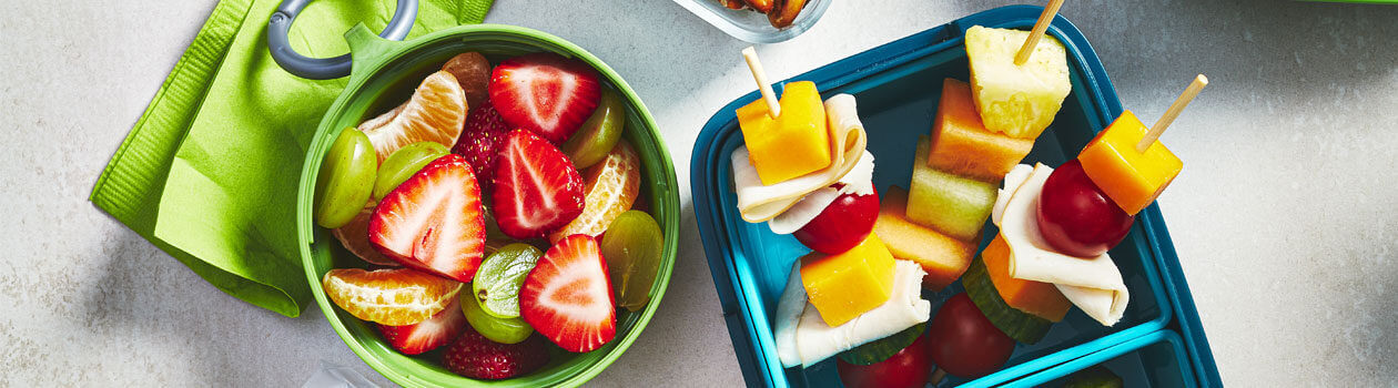 Mini pita sandwiches, mini pizzas, fruit and veggies in a lunchbox sitting next to school books and binders.