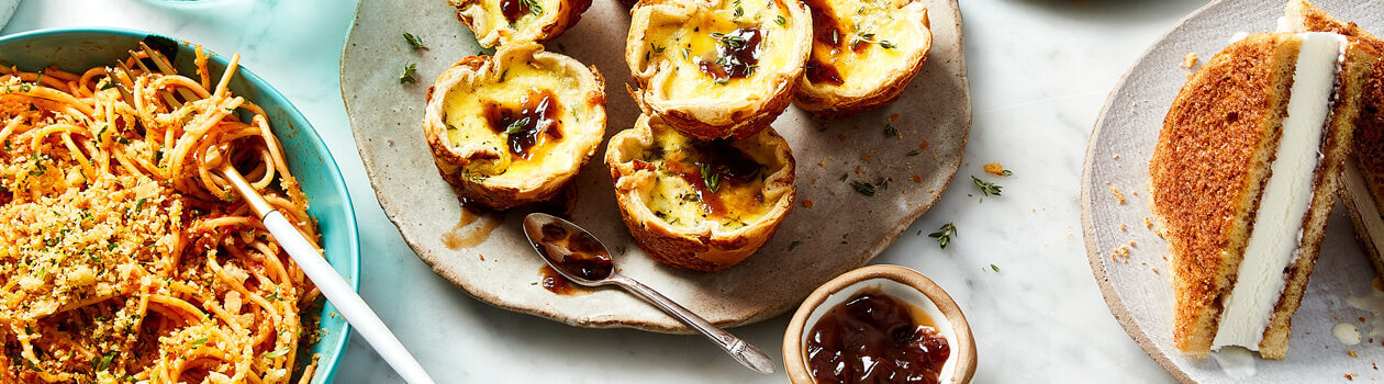 Hamburger bun mini quiches, cinnamon bread ice cream sandwiches and seasoned Italian-style breadcrumbs on top of a tomato sauced pasta in a bowl over a marble countertop.