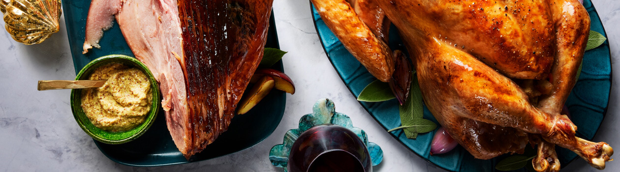 Roasted turkey and ham on blue platters sitting atop a marble counter with a side dish of roasted vegetables.