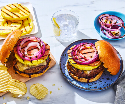 Two plates, each with Hawaiian-style burger topped with grilled pineapple round and side bowl of ruffled plain potato chips