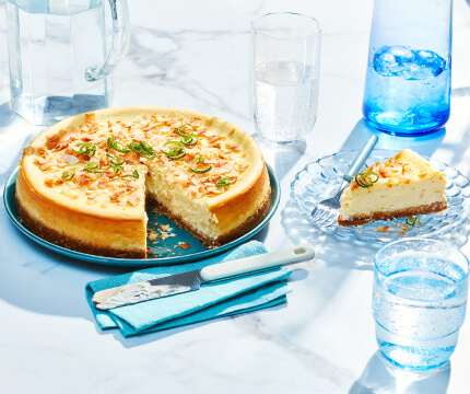 Full lime coconut cheesecake with lime peel garnish on serving plate with one slice removed and on plate to the right side.