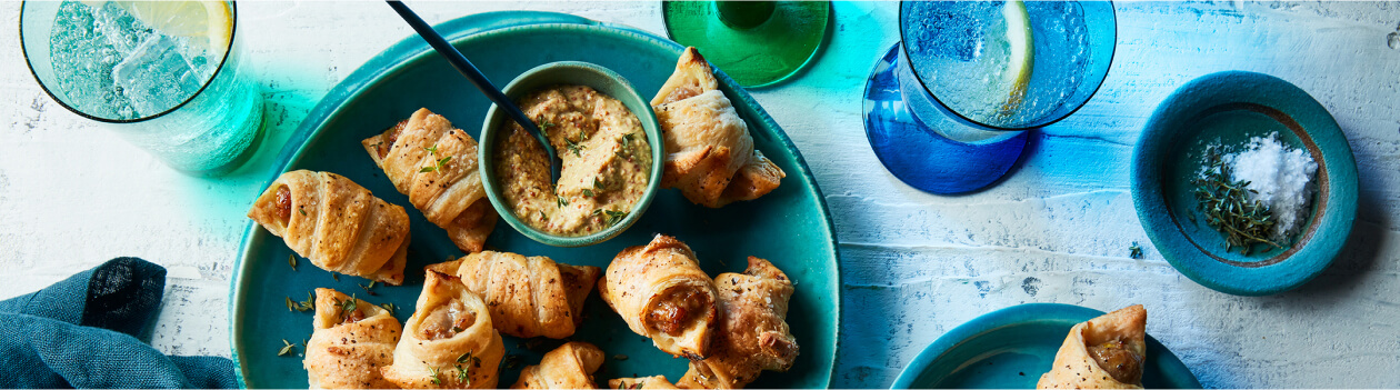 Tablescape with sparkling waters, napkin and round aqua green-blue serving plate with bratwurst pigs in blankets and a small bowl of grainy Dijon mustard for serving