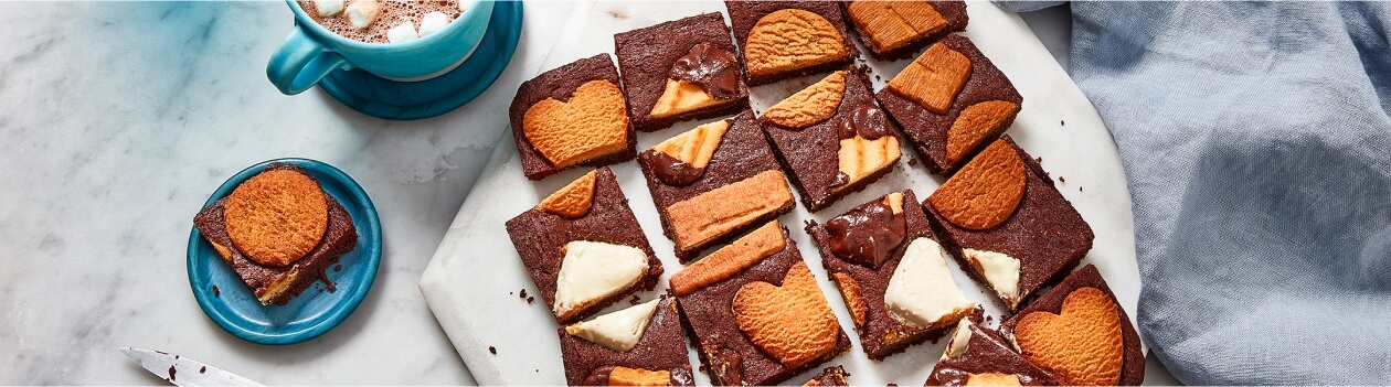 White marble surface with white cutting board of cut up brownies, studded across the top with Panache Belgian Biscuit cookie assortment