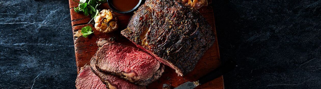 Roasted Sterling Silver prime rib on a wooden cutting board next to a roasted head of garlic with a carving knife.