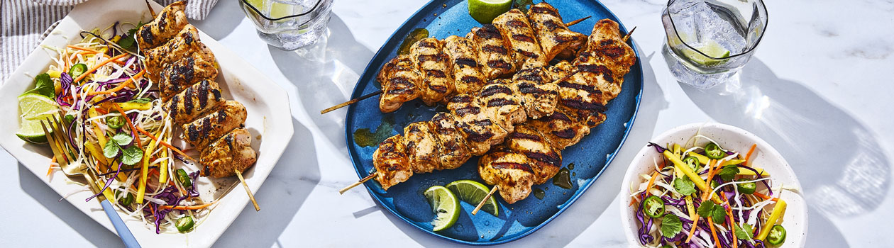 blue serving platter of Spicy Grilled Chicken Kabobs with Mango Slaw and single serve plate of kabobs and salad.