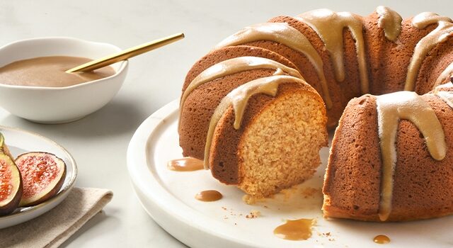 A bundt cake with a stick toffee glaze and fresh figs on top, a bowl of the toffee glaze, and a slice on another plate with a fork and a couple pieces of fig.