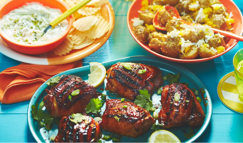 Blue wooden surface with plate of grilled chicken, bowl of ruffled potato chips and a side potato salad