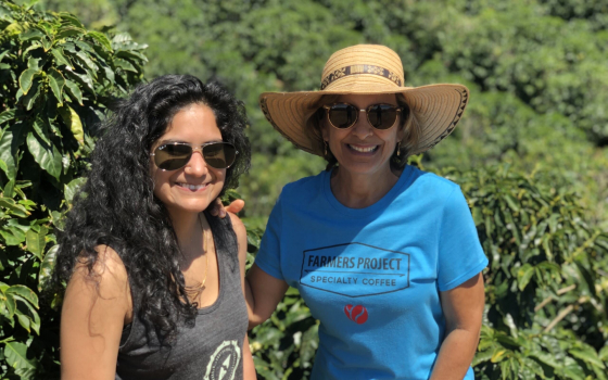 One woman is in a blue t-shirt with her hand resting on another woman’s shoulder, and she is in a grey tank top with vegetation behind them.