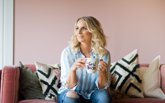 A blonde woman in a blue and white dress shirt sitting on a pink couch with a mug in her hands that says Girl Boss.