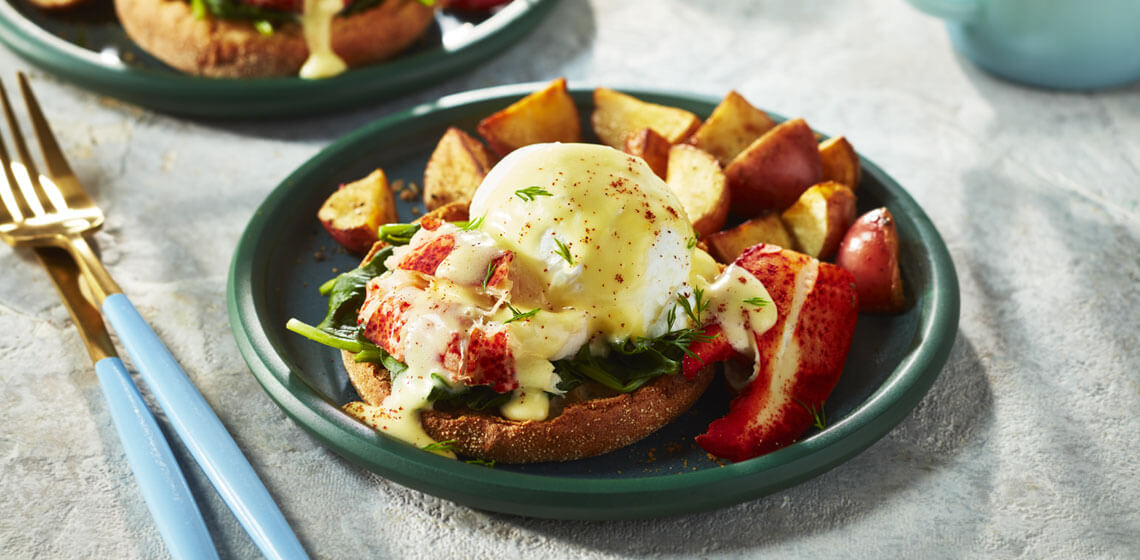 Eggs Benny topped with hollandaise sauce and a piece of steamed lobster next to home fries on a dark green plate.