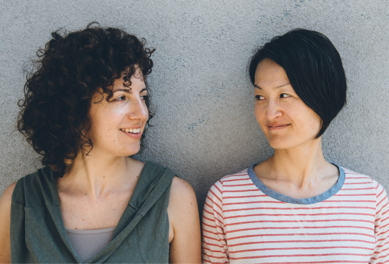 Two women looking at each other with a grey wall behind them.