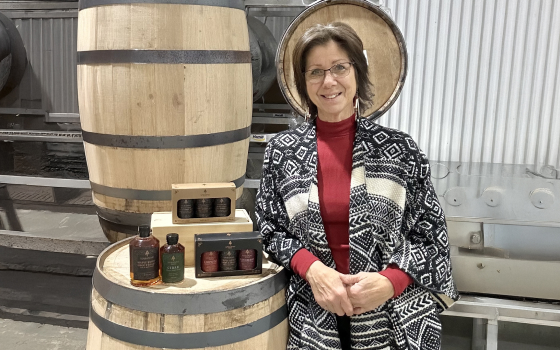 A woman leaning against a barrel with bottles of maple syrup on top.