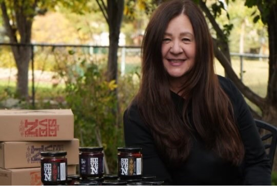 A woman dressed in all black, sitting at a table outside with a number of jarred oils stacked on top of it.