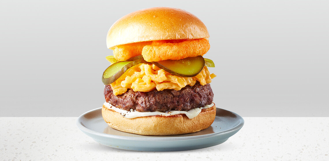 Compliments burger topped with Compliments macaroni and cheese, cheese puffs and bread and butter pickles on a blue/gray plate.