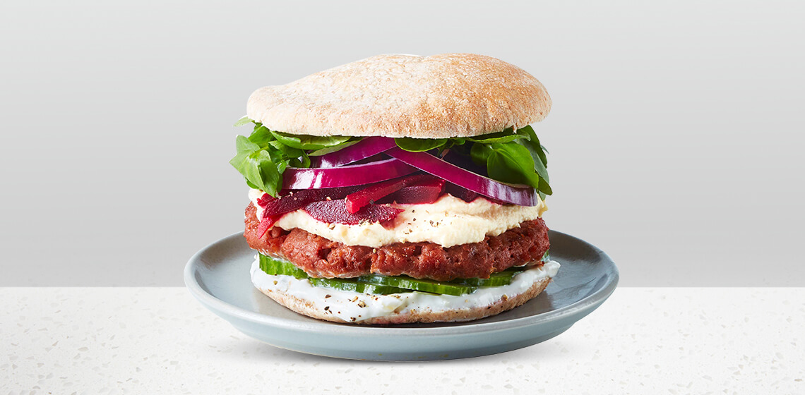 Beyond Meat burger patty on a whole wheat pita, topped with hummus, pickled beets, cucumber slices, tahini, red onion and arugula on a blue gray plate.