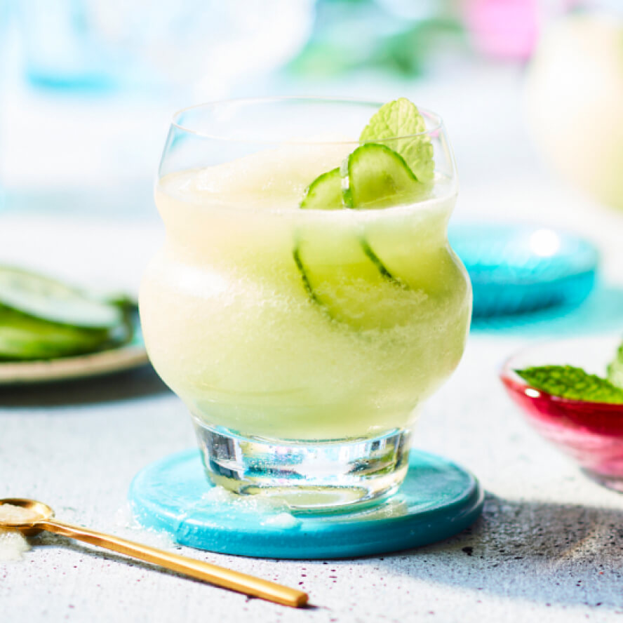 Lowball glasses of pale-green Cucumber Gimlet Slush sitting on top of a turquoise coaster on a grey countertop.